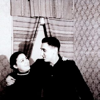 Two people posing affectionately indoors, mid-20th century.