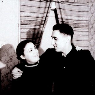 Two people posing affectionately indoors, mid-20th century.