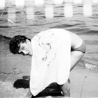 Man kneeling on beach, shirt stained, near water.