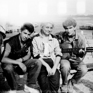 Three people posing outdoors with vintage guitar, 1970s-style clothing and industrial background.
