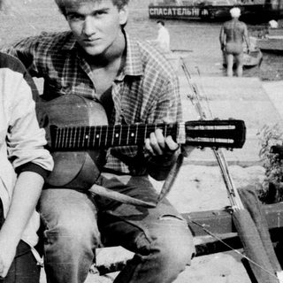 Three people posing outdoors with vintage guitar, 1970s-style clothing and industrial background.