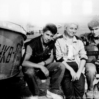 Three people posing outdoors with vintage guitar, 1970s-style clothing and industrial background.