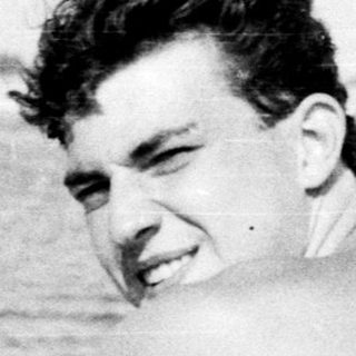 Young man sitting shirtless on beach towel.
