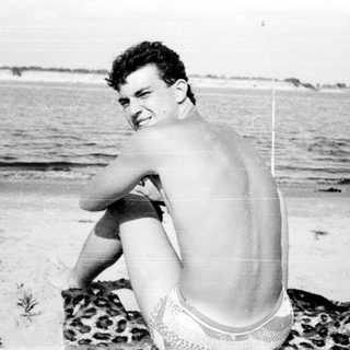 Young man sitting shirtless on beach towel.