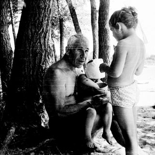 Shirtless man feeding young girl outdoors near trees.