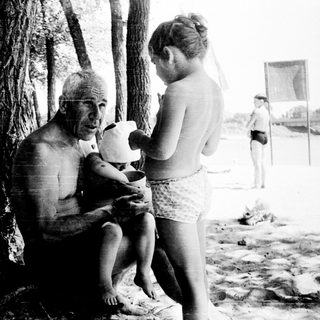 Shirtless man feeding young girl outdoors near trees.