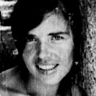 Shirtless person smiling behind tree trunk.