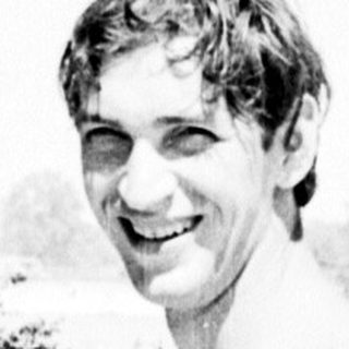 Shirtless man smiling outdoors near water.
