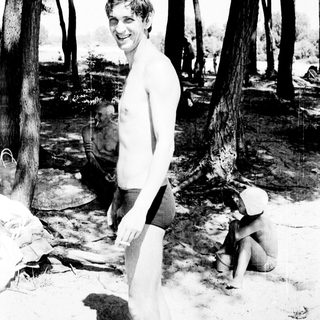 Shirtless man smiling outdoors near water.