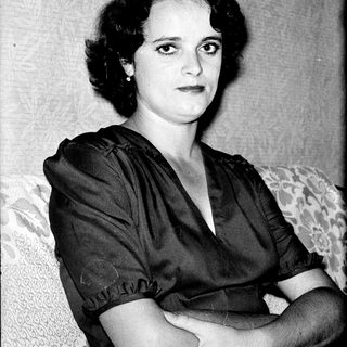 Woman in 1940s-style dress, seated with crossed arms.