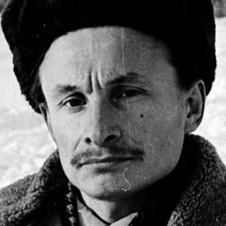 Man in winter attire sitting outdoors on snowy ground.