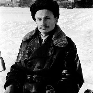 Man in winter attire sitting outdoors on snowy ground.