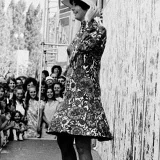 Woman in vintage dress speaking to crowd near tall wall.