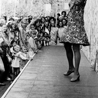 Woman in vintage dress speaking to crowd near tall wall.