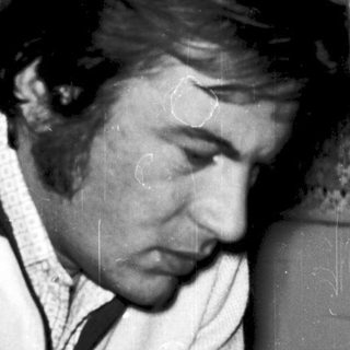 Man in 1970s jacket using flip phone indoors.