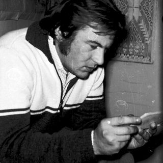 Man in 1970s jacket using flip phone indoors.