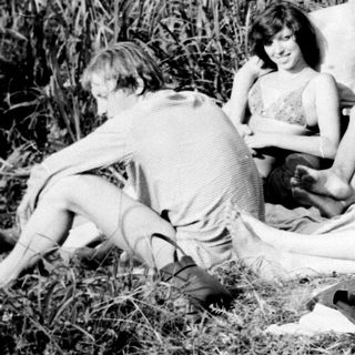 Group of people picnicking by water, dressed in 1970s-style swimwear.