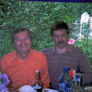 Two men posing outdoors at a garden table with drinks.