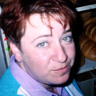 Woman in bakery wearing apron, surrounded by baked goods.