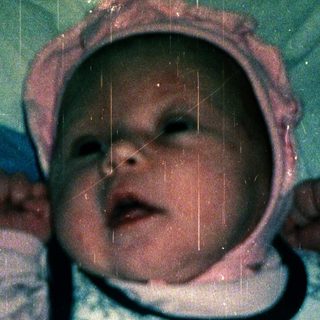 Baby wearing pink hat, lying on patterned blanket.
