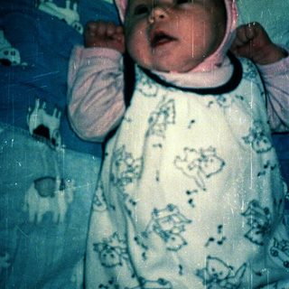 Baby wearing pink hat, lying on patterned blanket.