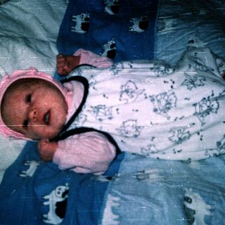 Baby wearing blue and white onesie, swaddled with pink hat, lying down.