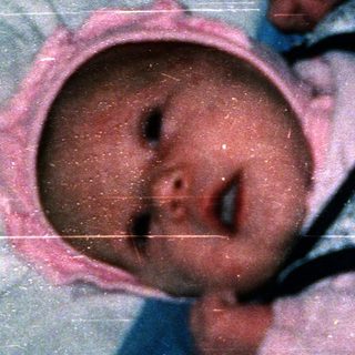 Baby wearing blue and white onesie, swaddled with pink hat, lying down.