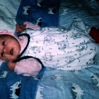 Baby wearing blue and white onesie, swaddled with pink hat, lying down.