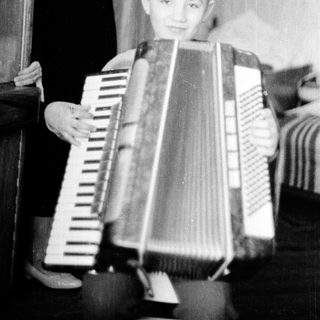 Young boy playing accordion indoors with adult nearby.