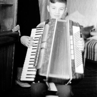 Young boy playing accordion indoors with adult nearby.