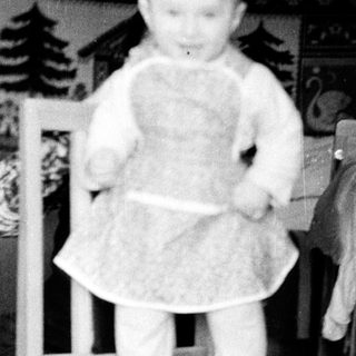 Baby standing on chair wearing dress.