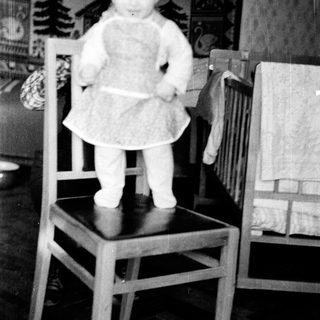 Baby standing on chair wearing dress.