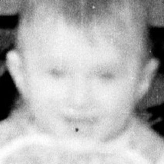 Baby standing on chair wearing dress.