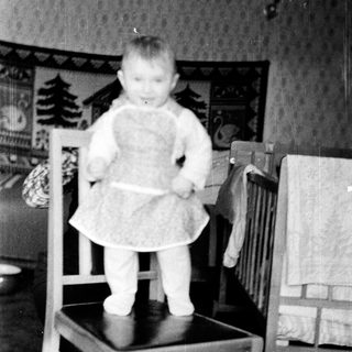 Baby standing on chair wearing dress.