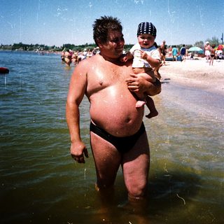 Man holding baby in shallow beach water.