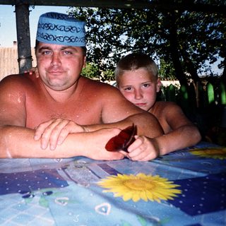Shirtless man and boy sitting outdoors by water.