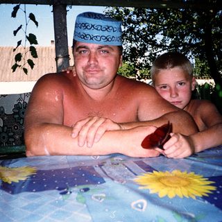 Shirtless man and boy sitting outdoors by water.