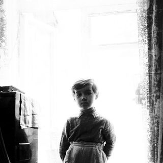 Young boy standing near a window in dimly lit room.