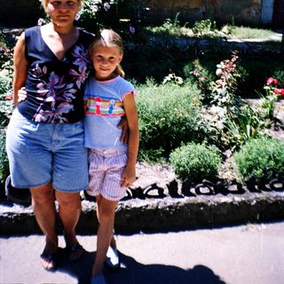 Two women posing outdoors near garden with stone structure.