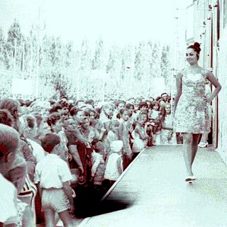 Woman in dress walking down crowded outdoor runway.
