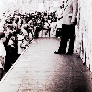 Man in suit addressing crowd at outdoor event.