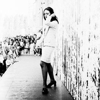 Woman in 1960s dress speaking into phone on city street.