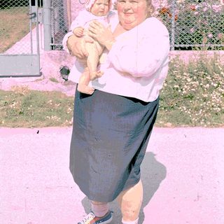 A woman holding a baby outdoors with another child in background.
