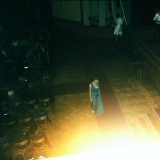 Person standing alone on stage in dimly lit hall.