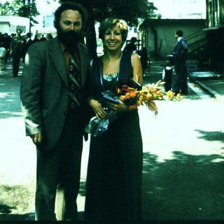 Two people holding floral offerings outdoors near trees.