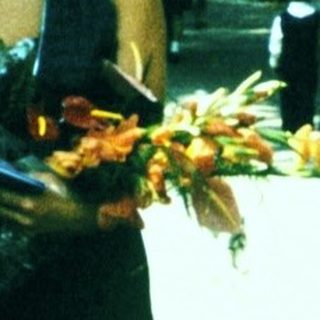 Two people holding floral offerings outdoors near trees.