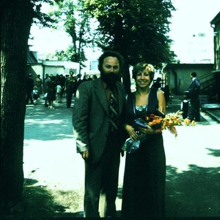 Two people holding floral offerings outdoors near trees.