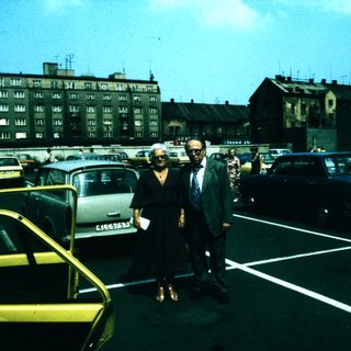 Two people standing in a mid-20th-century parking lot.