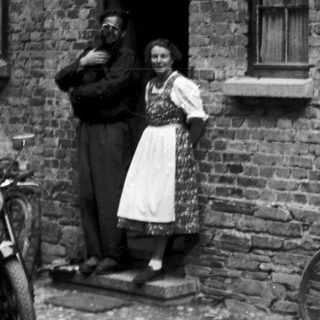 Vintage photo of couple standing by brick house with motorcycle and bicycle.
