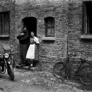 Vintage photo of couple standing by brick house with motorcycle and bicycle.